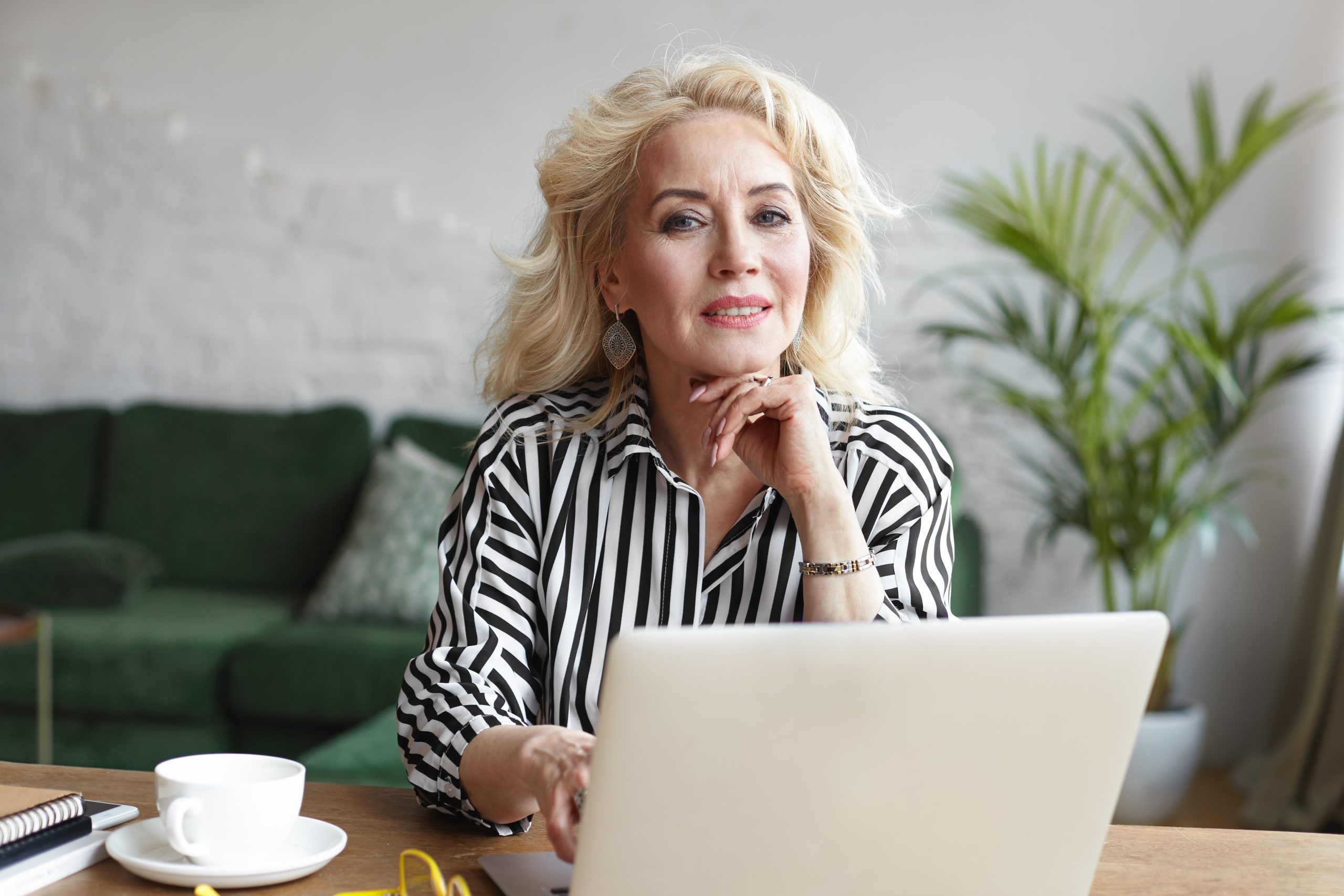 People, age, job and occupation concept. Attractive confident female on retirement wearing stylish striped blouse earning money via internet, using laptop for distant work from home office